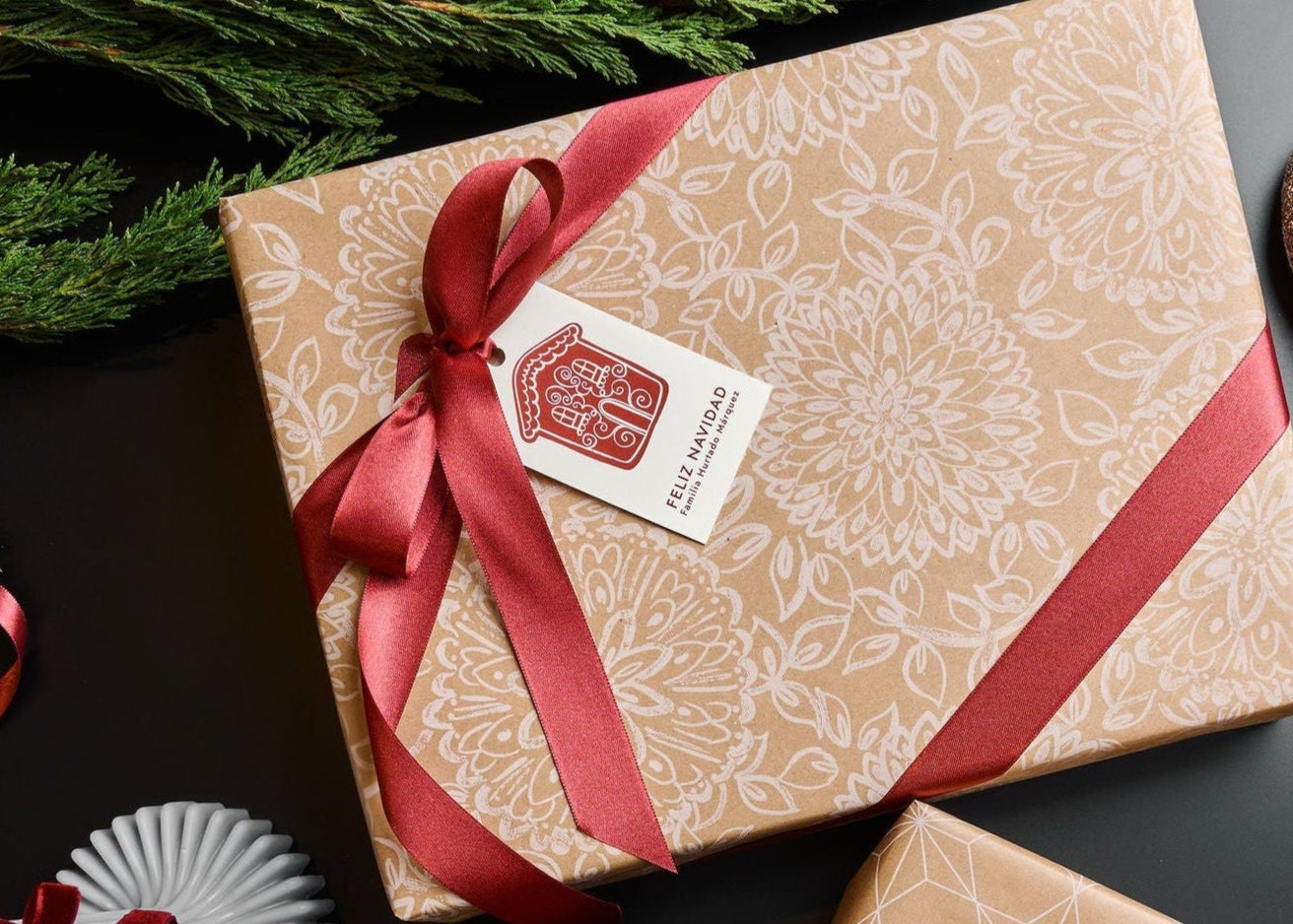 Two gift boxes with red ribbons and decorative tags on a dark surface with Christmas decorations.